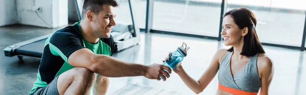 Plano panorámico de deportista sonriente dando botella deportiva a deportista en centro deportivo - foto de stock