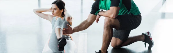 Panoramic shot of sportswoman doing crunches and sportsman helping her in sports center — Stock Photo
