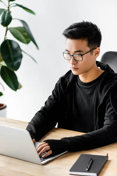 Asiatischer Hacker mit Brille sitzt am Tisch und benutzt Laptop — Stockfoto