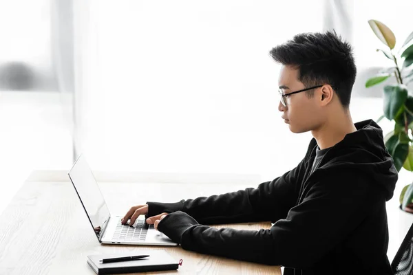 Vista lateral de hacker asiático en gafas sentado en la mesa y el uso de ordenador portátil - foto de stock