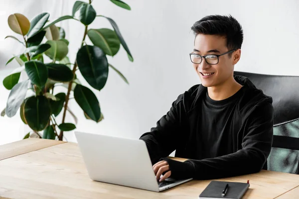 Улыбающийся азиатский хакер в очках сидит за столом и использует ноутбук — стоковое фото