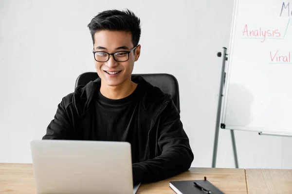 Smiling seo manager in glasses sitting at table and using laptop — Stock Photo