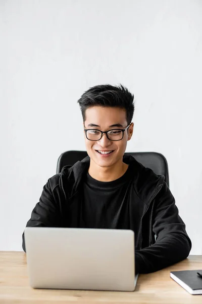 Улыбающийся азиатский хакер в очках сидит за столом и использует ноутбук — стоковое фото