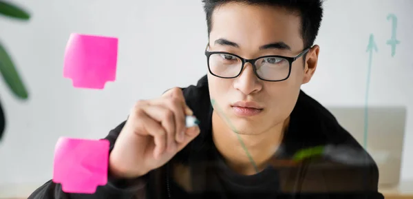 Panoramaaufnahme eines asiatischen SEO-Managers in einer Brille, der Pfeil auf Glas malt — Stockfoto