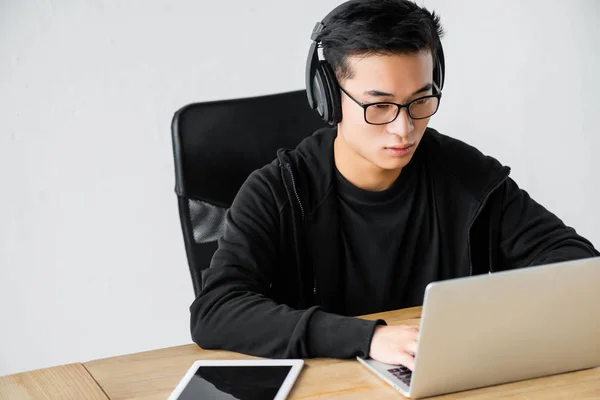 Азіатський хакер в навушниках, використовуючи ноутбук і сидячи за столом — стокове фото