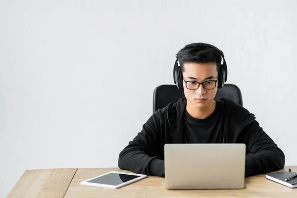 Hacker asiático en los auriculares usando el ordenador portátil y sentado en la mesa - foto de stock