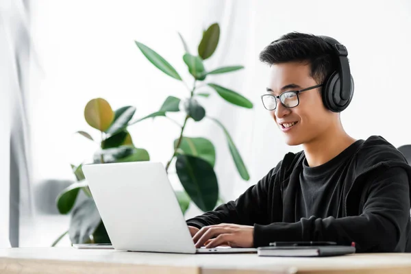 Smiling asian hacker in headphones using laptop and sitting at table — Stock Photo