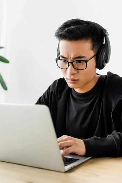 Asian hacker in headphones using laptop and sitting at table — Stock Photo