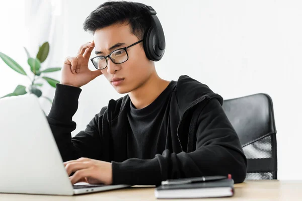 Thoughtful asian hacker in headphones using laptop and sitting at table — Stock Photo