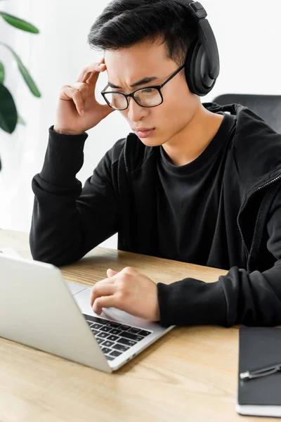 Vista de ángulo alto de hacker asiático reflexivo en los auriculares con el ordenador portátil y sentado en la mesa - foto de stock