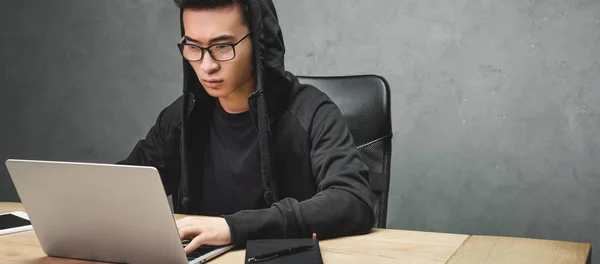 Panoramaaufnahme eines asiatischen Hackers, der mit Laptop am Tisch sitzt — Stockfoto