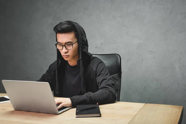 Hacker asiático en gafas usando portátil y sentado en la mesa - foto de stock