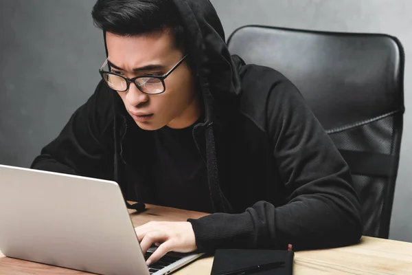 Hacker asiático en gafas usando portátil y sentado en la mesa - foto de stock