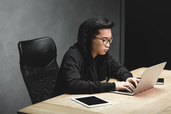 Hacker asiático en gafas usando portátil y sentado en la mesa - foto de stock