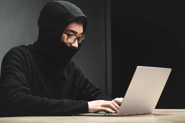 Азиатский хакер с затемненным лицом с помощью ноутбука и сидя за столом — стоковое фото