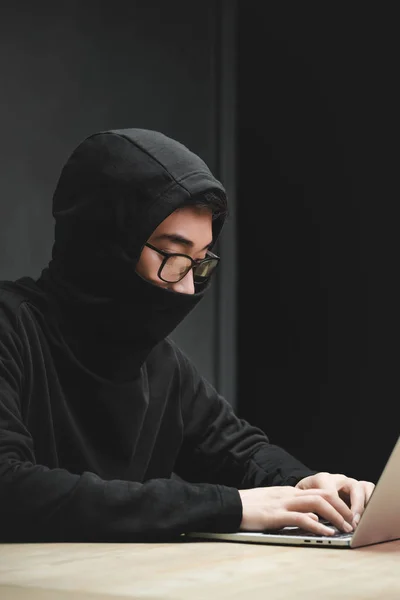 Азиатский хакер с затемненным лицом с помощью ноутбука и сидя за столом — стоковое фото