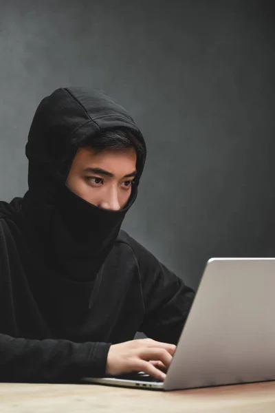 Hacker asiático con la cara oscurecida usando el ordenador portátil y sentado en la mesa - foto de stock