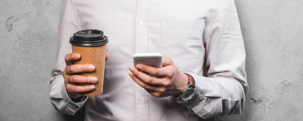 Plano panorámico de hombre de negocios sosteniendo taza de papel y el uso de teléfono inteligente - foto de stock