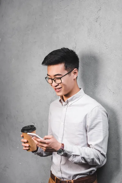 Улыбающийся азиатский бизнесмен держит бумажную чашку и использует смартфон — стоковое фото