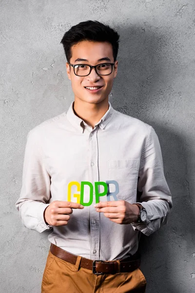 Улыбающийся азиатский бизнесмен, держащий красочные буквы gdpr и смотрящий в камеру — стоковое фото