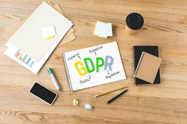 Top view of paper with gdpr lettering, notebooks, paper cup, pen, pencil, sticky notes, marker, smartphone and crumpled papers on wooden table — Stock Photo