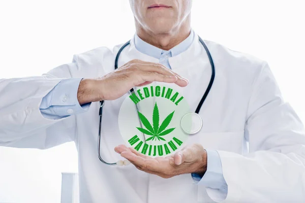 Vista ritagliata del medico in mantello bianco tenendo le mani intorno illustrazione marijuana medicinale isolato su bianco — Foto stock