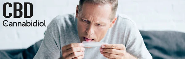 Panoramic shot of handsome man licking paper for blunt with medical cannabis near cbd lettering — Stock Photo
