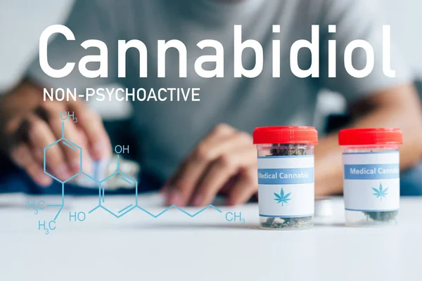 Selektiver Fokus von Flaschen mit medizinischem Cannabis auf dem Tisch mit Mann im Hintergrund mit nicht-psychoaktiver cbd-Illustration — Stockfoto