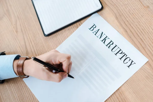 Vista superior del hombre sosteniendo la pluma cerca del papel con cartas de bancarrota y cuaderno - foto de stock