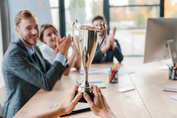 Cropped view of businesswoman holding champion cup near coworkers in office — Stock Photo