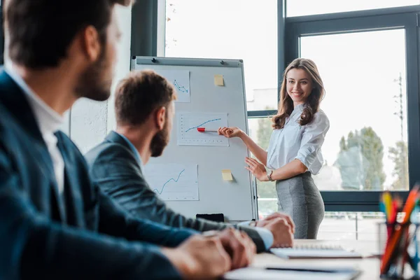 Selective focus of beautiful speaker smiling while looking at businessmen in office — Stock Photo