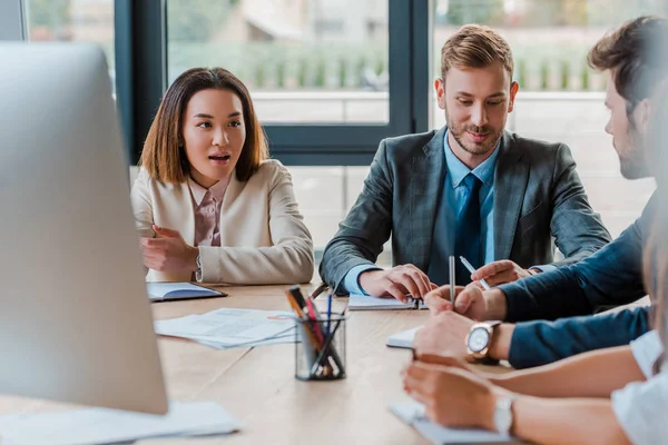 Enfoque selectivo de asiático mujer de negocios sentado con guapo hombre de negocios mientras mira a compañeros de trabajo - foto de stock