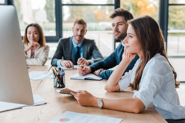 Selective focus of attractive businesswoman looking at computer during webinar monitor near multicultural coworkers in office — Stock Photo