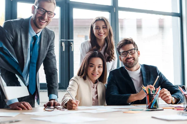 Foyer sélectif des hommes d'affaires heureux et femme d'affaires multiculturelle regardant la caméra dans le bureau — Photo de stock