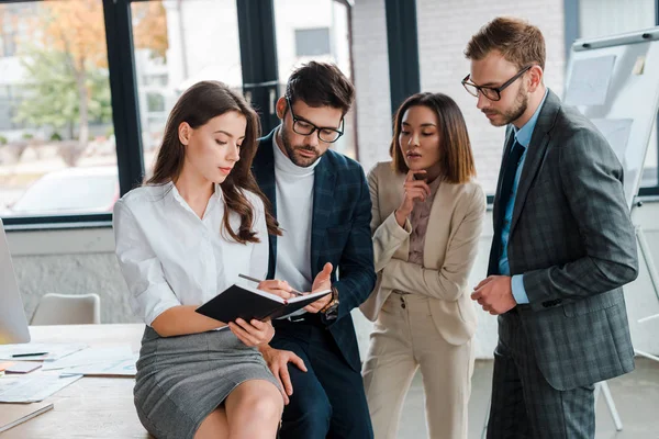 Handsome businessmen in formal wear standing near attractive multicultural businesswomen in office — Stock Photo