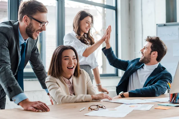 Selective focus of happy businessman in glasses smiling near multicultural coworkers giving high five in office — Stock Photo