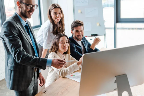 Hombre de negocios feliz en gafas que sostienen la pluma y mirando el monitor de computadora cerca de compañeros multiculturales en la oficina - foto de stock