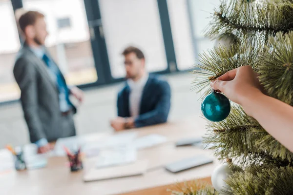 Vista recortada de la mujer tocando la bola de Navidad en el árbol de Navidad cerca de los hombres de negocios en la oficina - foto de stock