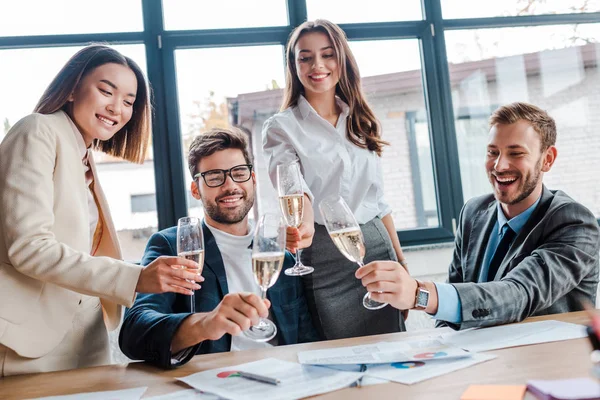 Cheerful multicultural businesswomen and businessmen holding champagne glasses in office — Stock Photo