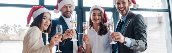 Panoramic shot of happy multicultural businesswomen and businessmen in santa hats holding champagne glasses in office — Stock Photo