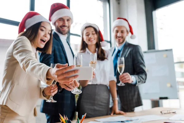 Foco seletivo de empresários multiculturais felizes e empresários em chapéus de santa tirar selfie enquanto segurando copos de champanhe no escritório — Fotografia de Stock