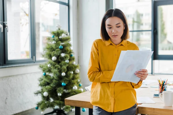 Attraktive asiatische Geschäftsfrau mit Diagrammen und Diagrammen in der Nähe des geschmückten Weihnachtsbaums — Stockfoto