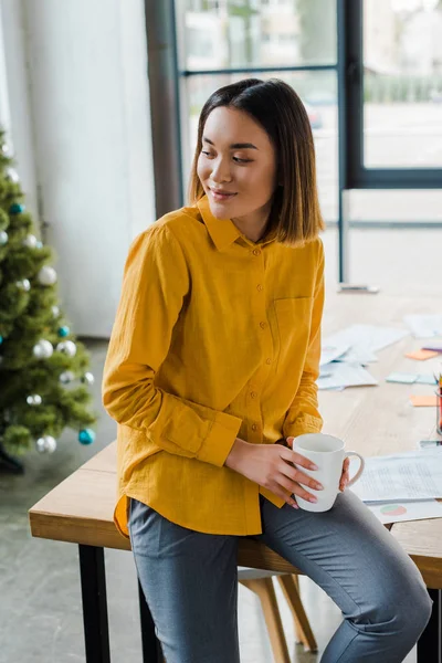 Cheerful asian woman holding cup with coffee near decorated christmas tree — Stock Photo