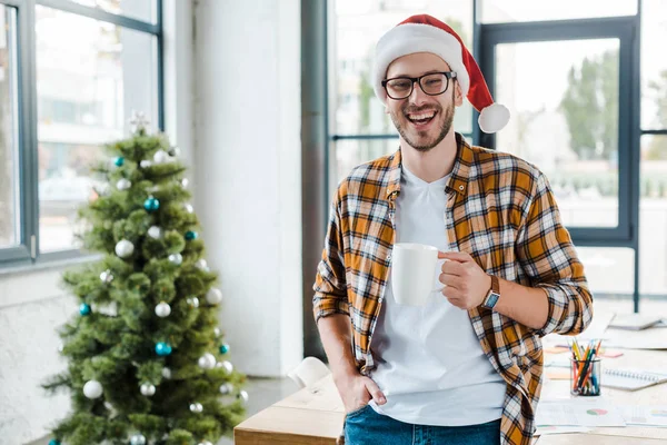 Messa a fuoco selettiva di allegro uomo barbuto in cappello Santa tenendo tazza vicino albero di Natale in ufficio — Foto stock