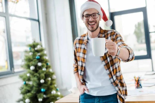 Selektiver Fokus eines glücklichen bärtigen Mannes mit Weihnachtsmannhut, der Tasse in der Nähe des Weihnachtsbaums im Büro hält — Stockfoto