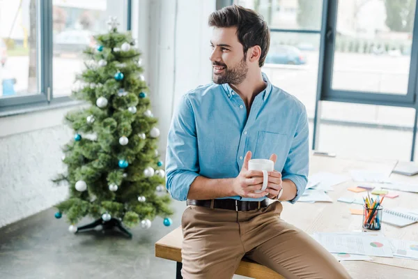 Enfoque selectivo del hombre barbudo positivo sosteniendo la taza cerca del árbol de Navidad en la oficina - foto de stock