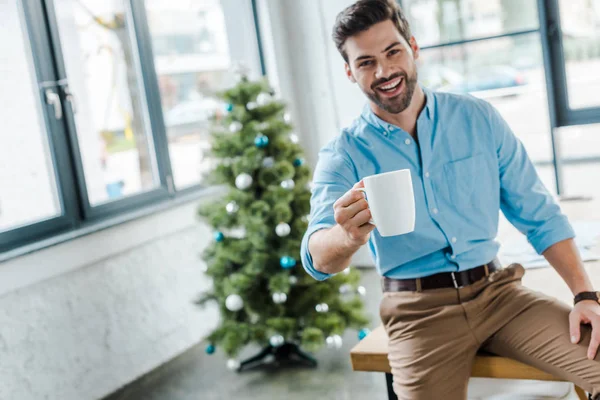 Вибірковий фокус щасливого бородатого чоловіка тримає чашку з кавою біля ялинки в офісі — стокове фото