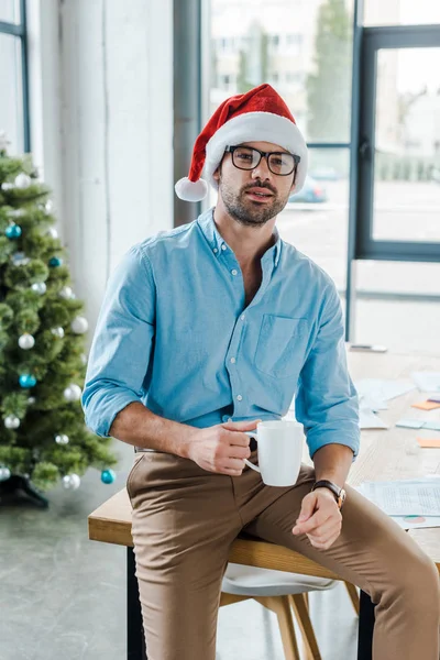 Selective focus of bearded man in santa hat and glasses holding cup with coffee near christmas tree in office — Stock Photo