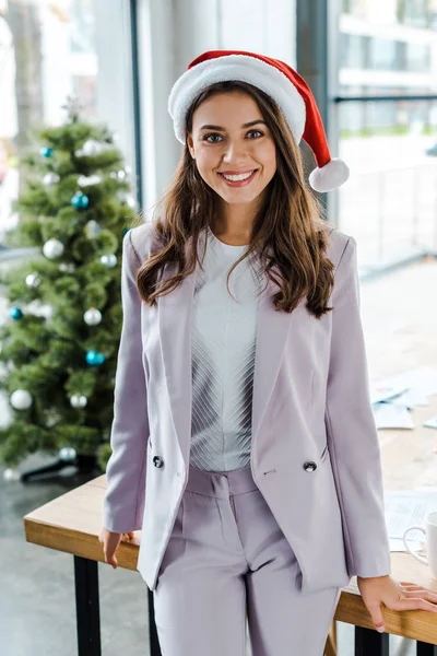 Cheerful businesswoman in santa hat near christmas tree in office — Stock Photo