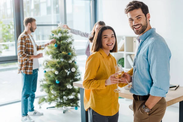 Enfoque selectivo de hombre de negocios y mujer de negocios asiática con copas de champán cerca de colegas y árbol de Navidad - foto de stock